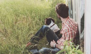 dog and man sitting in tall grass and leaning against a building